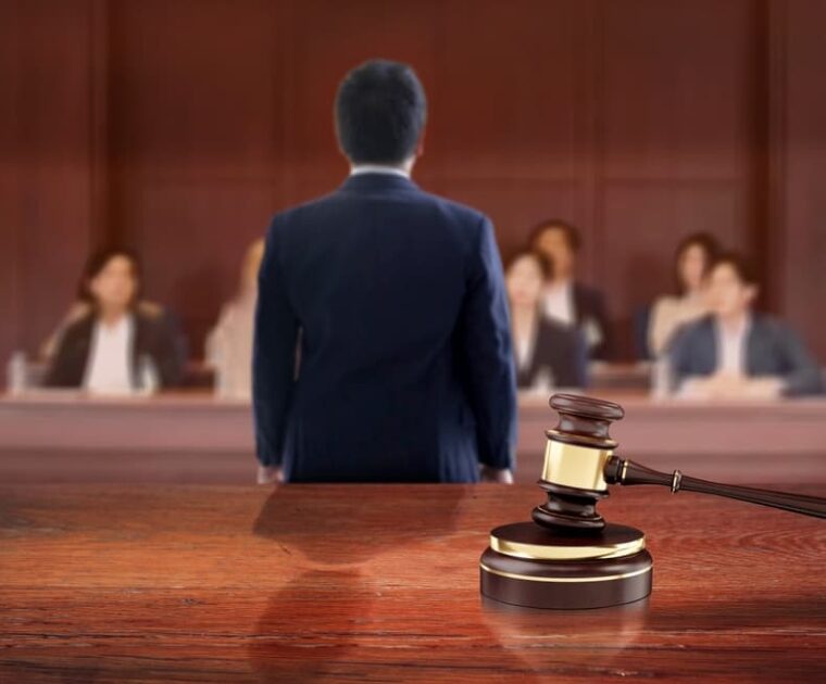 The judge's gavel symbolizes judgment as it rests on the judicial wooden table in the courtroom, while the lawyer addresses the jury in the background.