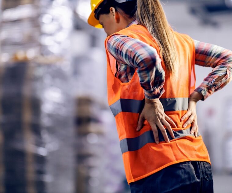 A female storage worker, seen from the back, massages her lower back, showing clear discomfort and pain from strenuous work.