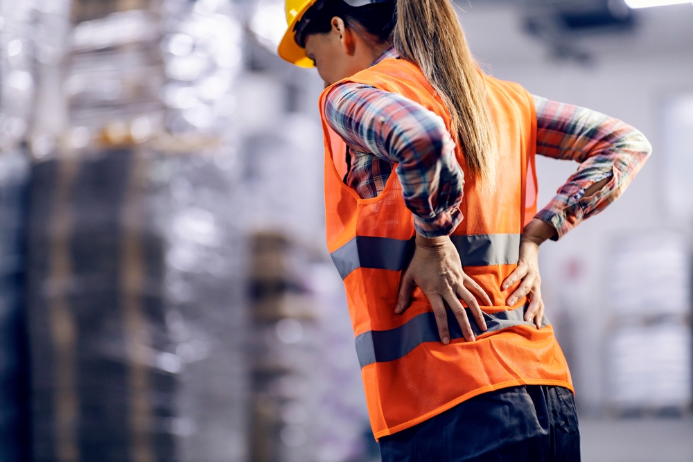 A female storage worker, seen from the back, massages her lower back, showing clear discomfort and pain from strenuous work.